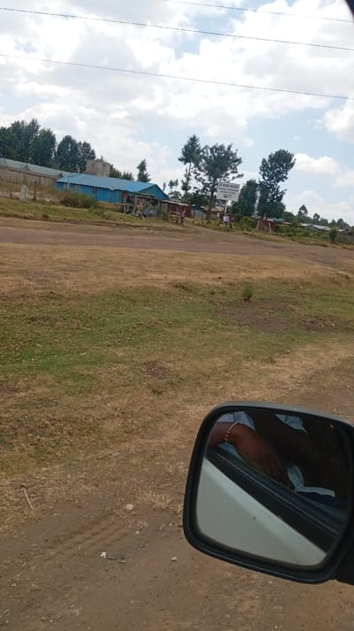 A landscape view from a car window features dry grassy terrain leading to a cluster of buildings. The closest building is blue with a corrugated roof. There is a sign reading "Lee Niiru Resort." Trees line the horizon, and a cloudy sky fills the upper portion of the frame. A side mirror is in the foreground, showing a partial reflection of an arm. The ground near the car shows dirt and tire tracks.
