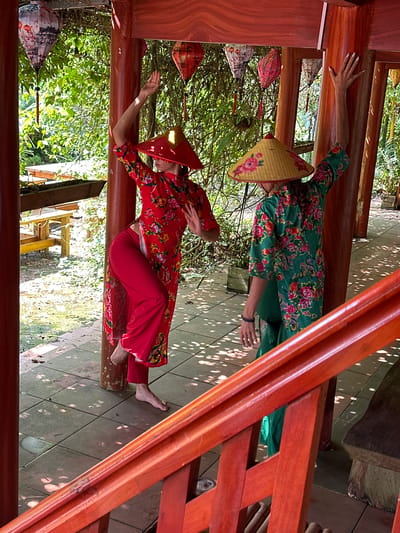 Two individuals, likely women, are captured in a bright, sunlit outdoor setting. They are both barefoot and adorned in traditional Vietnamese-style clothing featuring vibrant floral patterns. The person on the left wears a red patterned outfit with red trousers and a matching red conical hat (nón lá). Her right hand is placed on a vertical wooden beam, and her left arm is bent with her hand near her chest, suggesting a posed or dance-like movement. The person on the right wears a green patterned outfit and a light yellow or beige conical hat, with her left hand raised against a horizontal wooden beam.

The scene takes place within a wooden structure, possibly a traditional Vietnamese house or pavilion, characterized by sturdy wooden pillars and railings. Beyond the structure, lush green foliage and trees are visible, indicating an abundant natural environment. Several decorative lanterns of various colors and patterns hang suspended, adding to the cultural ambiance. The ground is a tiled floor, upon which patches of dappled sunlight create bright, dynamic patterns, suggesting it is a sunny day, likely mid-morning or early afternoon. There is no visible text in the image. The overall impression is one of graceful movement in a serene, culturally rich outdoor setting in Vietnam.