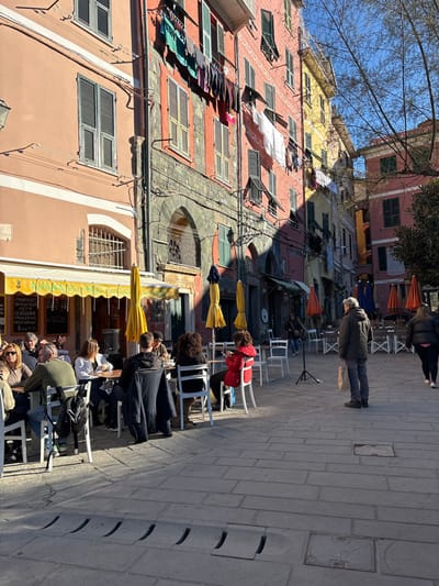 This image captures a sunlit street scene in Vernazza, Italy. The view is down a pedestrian thoroughfare flanked by colorful, multi-story buildings, characteristic of Ligurian architecture. Buildings display shades of peach, red, and yellow, featuring numerous windows fitted with louvered shutters, some grey and others green. Laundry, including assorted garments, hangs from lines strung between and from the upper floors of the buildings.
In the left foreground, an outdoor cafe is active, identified by its bright yellow awnings and matching stand-alone umbrellas. Approximately ten individuals are seated at white tables with white chairs, engaged in various interactions such as conversing, eating, and drinking. A dark backpack rests on one of the chairs. Menu boards are visible beneath the cafe awning.
The central and right portions of the paved pedestrian area feature fewer occupants. A male individual with grey hair, dressed in a dark jacket and blue jeans, stands facing right, holding a light-colored bag. He is positioned as if observing the scene. Further right, a female individual in a dark jacket is walking away from the viewer. Several white chairs and small tables are arranged in the midground and background, some unoccupied, alongside closed orange umbrellas. A black tripod stand, devoid of mounted equipment, is centrally located on the paving. Strong sunlight from a clear sky casts distinct shadows across the street and the lower portions of the buildings. Bare tree branches are visible against the blue sky on the upper right. The ground is paved with rectangular stone blocks, and a linear drainage grate with curved slots is present in the immediate foreground.