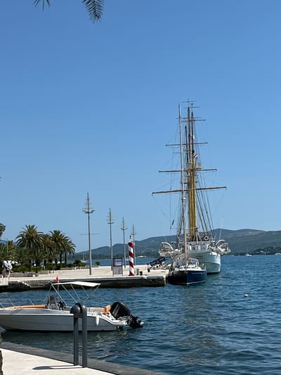 The image shows a waterfront scene in Donja Lastva, Montenegro, on a clear, sunny day. A large sailing ship, possibly a training vessel, is docked at a pier. The ship has multiple masts and rigging, with the name "JADRAN" visible on its stern. Moored alongside the sailing ship is a smaller, dark-hulled boat. In the foreground, a white speedboat with an orange interior and a black outboard motor is tied to a dock. The water is a deep blue, with ripples indicating a light breeze.
Behind the pier, a walkway lined with palm trees leads to buildings. Several tall, decorative lampposts with ship-like tops are also visible along the waterfront. A red and white striped lighthouse stands on the pier, near the sailing ship. In the distance, a hilly, tree-covered landscape forms the background, with more boats scattered across the sea. The sky is a clear, bright blue. No people are clearly visible interacting with the boats or the environment.