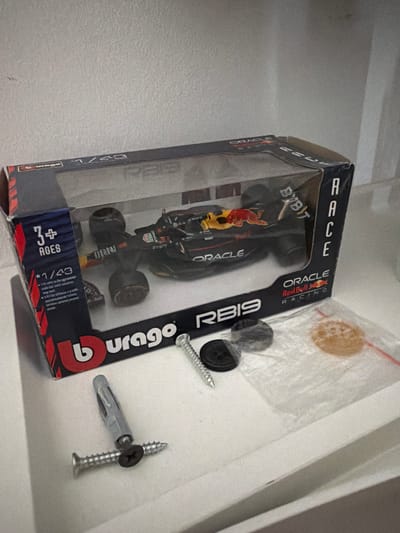 The image shows a Burago 1/43 scale model car of the Oracle Red Bull Racing RB19. The box indicates that the toy is suitable for ages 3+. The model is still within its packaging. Adjacent to the box are screws and plastic wall anchors. The photo appears to be taken indoors, likely inside a home or storage area. There is no visible indication of Pétange, Luxembourg in this image.