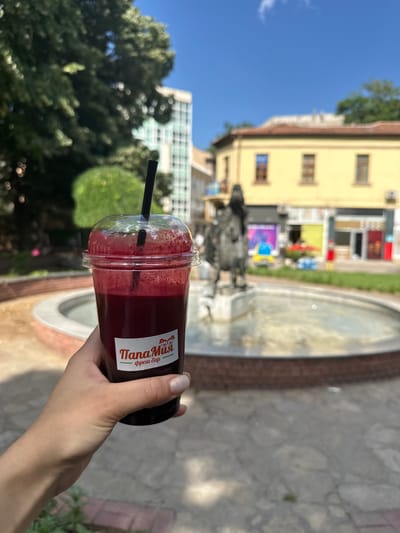 A person's hand, with neatly manicured nails, holds a clear plastic cup filled with a dark red liquid, likely a juice or smoothie, topped with a clear plastic lid and a black straw. A sticker on the cup features a logo with two cartoon bears and text that reads "ПапаМия" and "фреш бар". The background is blurred but suggests an outdoor urban setting with a fountain in the foreground, a statue within the fountain, and buildings behind it. The architecture of the buildings, particularly the yellow one with arched windows and a red tile roof, is characteristic of older European city centers. The presence of a modern glass-fronted building to the left of the yellow building indicates a mix of architectural styles. The sky is clear and blue. This scene is located in the central square of Haskovo, Bulgaria, specifically near the fountain and the iconic building of the former Municipal Bank, which now houses shops and cafes.