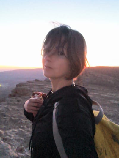 A young woman with dark, shaggy hair stands on a rocky, elevated surface, possibly a ridge or cliff, overlooking a valley bathed in the soft light of dusk or dawn. Her gaze is directed slightly away from the camera, and she holds a piece of food, appearing to be a sandwich or pastry, in her left hand, bringing it towards her mouth. She is wearing a dark hooded jacket, and a yellow backpack with gray straps is visible on her right side, partially obscured by her body. The background features layers of hills and a gradient sky transitioning from a light blue at the top to a soft orange glow near the horizon, suggesting a natural landscape on the outskirts of Yeruham, Israel, likely within the vicinity of the Yeruham Crater (Makhtesh Yeroham). The terrain is arid and rocky, characteristic of the Negev Desert.