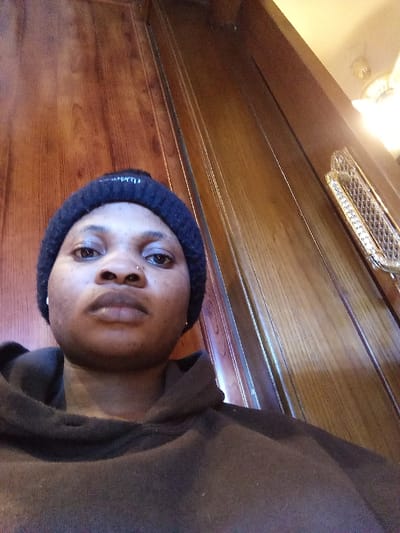 A close-up, slightly low-angle portrait shot depicts an individual with dark skin, wearing a dark blue knit beanie with a white embroidered logo and a dark-colored hooded sweatshirt. A small silver-toned stud piercing is visible on the subject's left nostril. The subject maintains a direct, neutral gaze towards the camera. The background consists of a vertical wooden paneling with a reddish-brown grain. To the right, a decorative gold-toned metallic fixture featuring an intricate diamond or lattice pattern is mounted on a wall, partially illuminated by an unseen light source. A cream-colored ceiling and a ceiling-mounted light fixture are visible in the upper right quadrant. The scene is an interior location in Madinaty, Egypt.