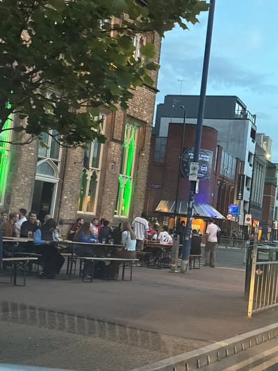 The image shows an outdoor seating area of a pub or bar in Manchester, United Kingdom. People are sitting at tables along the pavement. The architecture includes brick buildings with ornate windows highlighted by green lights, as well as more modern buildings in the background. There is a sign for "Frog & Bucket Comedy Club" with Pepsi branding visible. Street signs and traffic lights are also present. The people in the scene appear to be socializing, eating, and drinking.