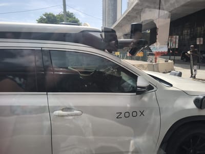 A white Zoox autonomous vehicle is parked on a street in Miami, Florida. The vehicle is a modified SUV with a sensor array mounted on its roof. The sensor array includes lidar, radar, and cameras. The vehicle is white and has black trim. The Zoox logo is visible on the side of the vehicle. A person is walking in the background with a crutch and a cast on their leg. The scene is a sunny day with blue skies. The background shows a cityscape with tall buildings and an overpass. The image is taken from the side of the vehicle, looking towards the front.