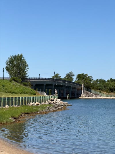 The image depicts a bright, clear day at a waterfront location, likely in the Village of Sag Harbor, United States of America. A large concrete bridge with railings and lampposts spans across a body of water. The bridge is supported by multiple concrete pillars. To the left of the bridge, a grassy hill is visible, adorned with a single, large, leafy tree and two lampposts. A wooden fence, painted green and weathered, runs along the edge of the land, separating it from the water. The shoreline is rocky with clusters of small to medium-sized rocks and patches of green grass growing near the water's edge. The water itself is a deep blue, with ripples on its surface reflecting the clear sky. On the right side of the bridge, the landscape transitions to a more densely wooded area with various trees. A sandy beach is present in the foreground on the left side of the image. There are no people or animals visible in the scene. The overall impression is one of a serene, natural landscape with a man-made structure integrated into it.