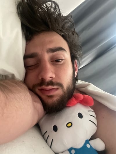 In a close-up, overhead shot, a young man lies in bed with his eyes closed, his head resting on a pillow. To his right, a plush Hello Kitty toy with a red bow rests against his cheek, its white face featuring black eyes, a yellow nose, and short black lines for whiskers. The man has dark, wavy hair, and a beard and mustache. He is wearing a blue shirt with white polka dots, and a white blanket is draped over him. The scene appears to be indoors, with gray curtains visible in the background. The lighting suggests it is daytime. The man's expression is relaxed, possibly indicating he is sleeping or resting. The overall mood is peaceful and intimate. The location context is Huixquilucan de Degollado, Mexico. There is no visible text in the image.