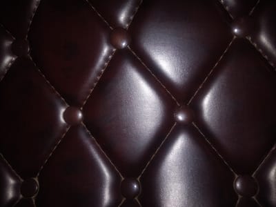 The image displays a close-up, textured view of dark brown, tufted upholstery, likely leather, characterized by diamond-shaped patterns formed by stitching and button accents. The lighting creates strong highlights and shadows, emphasizing the material's sheen and depth. There are no people or identifiable objects other than the upholstery itself. This image is not associated with any specific location in Haveli Jhangranwali, Pakistan, as it is a generic texture shot.