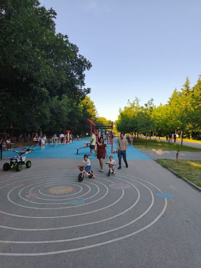 The image depicts a public park, possibly located in Haskovo, Bulgaria, with a playground and exercise area. In the foreground, a young girl on a pink balance bike rides on a circular path painted on the asphalt, which appears to be a solar system diagram. To her left, a white and green ATV-like ride-on toy is parked. Behind them, a man and a woman walk, with the man looking ahead and the woman glancing back.
The park features a vibrant blue surface where various exercise equipment, including pull-up bars and stationary machines, are set up. Several individuals are engaged in activities on these machines or are observing others. Lush green trees line the park, creating a shaded atmosphere, with the sun casting warm light on the scene. The sky is clear and blue, suggesting a pleasant day. In the background, more people are visible strolling along a pathway. The overall impression is a well-maintained, active community space.