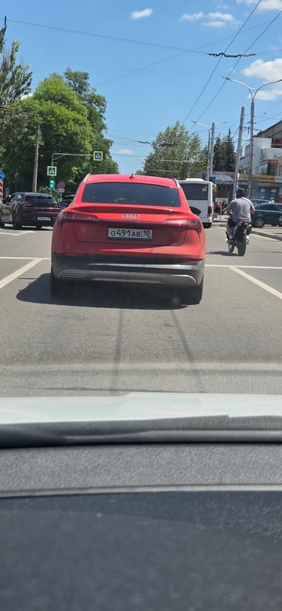 A red Audi SUV is stopped at a traffic light in Donetsk, Ukraine. The car's license plate reads "O491AB 10". A black motorcycle with a rider is also stopped at the light to the right of the Audi. In the background, other vehicles, including a white bus, and buildings are visible. The sky is blue with some clouds. The scene appears to be a typical city street on a sunny day.
