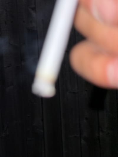 In Riverhead, United States of America, a close-up shot focuses on a white cylindrical object, possibly a cigarette, held by a blurred hand. The object is positioned in the foreground, with a dark, wooden fence serving as the background. A faint wisp of smoke is visible near the tip of the object. The lighting is dim, contributing to the overall blurred effect of the image, with the focus primarily on the object and the hand. The fence appears to be made of vertical wooden planks, suggesting an outdoor setting.