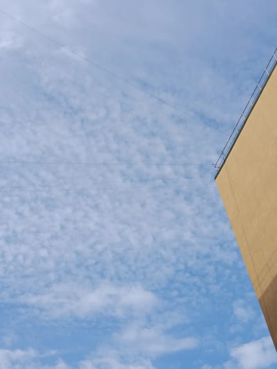 The image shows a corner of a light yellow building against a blue sky with scattered white clouds. Two thin dark lines, likely wires, are visible stretched horizontally across the sky. The building has a metallic railing along its upper edge. The scene is a close-up, abstract view of architecture and the sky. The image was taken in the city of Yablonovskiy, Russia.