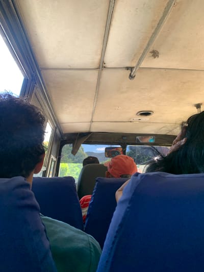 The image depicts the interior of a bus, viewed from the perspective of a passenger seated behind the driver and looking forward. The ceiling of the bus is white and appears worn, with metal supports visible. The windows offer a view of lush green trees and foliage, suggesting an outdoor environment.
In the foreground, the backs of two passengers are visible, one with dark, curly hair and wearing a blue shirt, and another wearing a green shirt. Further ahead, a passenger wearing an orange baseball cap is visible. The driver's area of the bus is partially obscured. The overall atmosphere suggests a journey through a rural or semi-rural area.
The question of the location in El Valle del Espíritu Santo, Venezuela, cannot be answered from the visual information provided in the image alone. There are no specific landmarks or textual cues within the image that can definitively place it within that city.