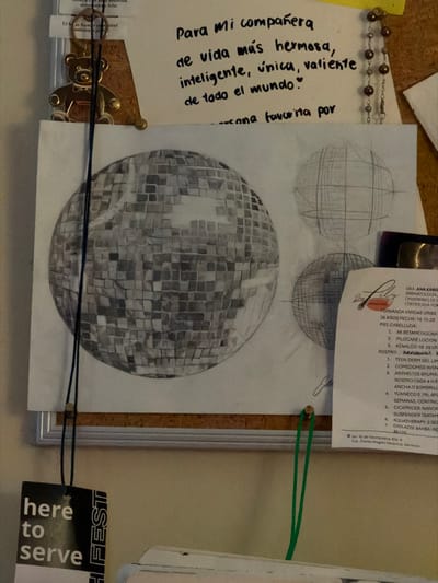 A corkboard displays various items. A sketch shows three stages of a disco ball. A medical prescription lists dermatology products. A handwritten note in Spanish expresses appreciation for a "beautiful, intelligent, unique, and brave" companion. Also present are a beaded chain and a tag with "here to serve" printed on it, attached with a black cord. The items are pinned to the board using push pins.