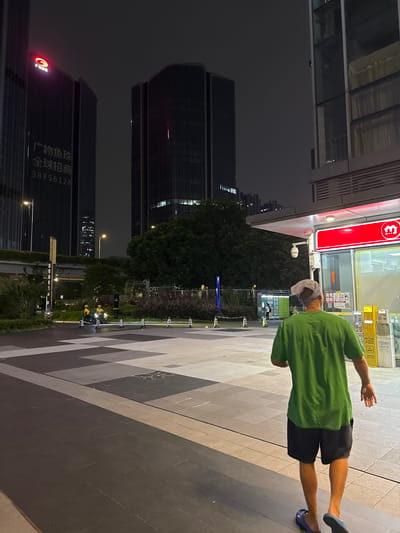 A man wearing a green t-shirt and dark shorts walks away from the camera at night on a city sidewalk in Huangpu District, China. He has a blue bandana on his head and is wearing blue slides. The sidewalk is paved with large tiles in a geometric pattern of grey, beige, and dark grey. In the background, there are modern skyscrapers with illuminated windows, and a green tree-lined area. To the right, there is a red and white convenience store with a M.V.I. logo and yellow vending machines. Two individuals on motorcycles wearing yellow uniforms are visible in the mid-ground. The overall scene is illuminated by artificial light from the buildings and store.