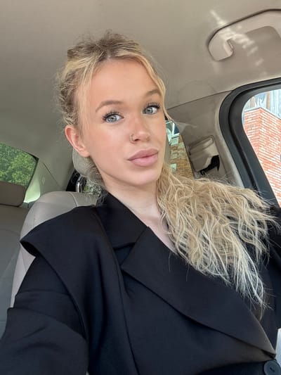 A blonde woman with blue eyes and a nose ring is taking a selfie in the passenger seat of a car. She is wearing a black blazer. The interior of the car is visible, including a beige seat and a grey headliner. Outside the car window, a brick building and some greenery can be seen. The image was taken in the city of Kommolovo, Russia.