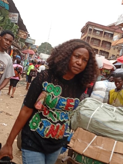 A woman in a black shirt with colorful lettering, carrying a handbag, stands in a bustling outdoor market scene. The background is filled with people, market stalls, and buildings. A young man is seen to the left, while a cart laden with packages is in the foreground to the right. The scene is busy and appears to be in a populated urban area.