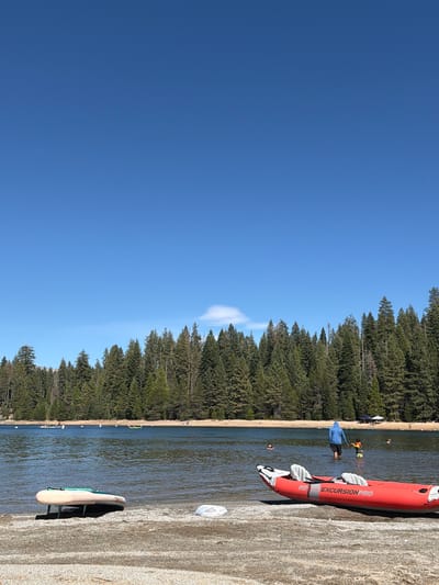 A bright, sunny day is depicted with a clear blue sky. The scene overlooks a calm body of water, likely a lake, bordered by a dense forest of evergreen trees. In the foreground, on a sandy and pebbly shore, rests a red inflatable kayak with the branding "EXCURSION PRO". To the left of the kayak, on its side, is a white paddleboard. Several people are visible in the water, some swimming, and two are standing near the shore. One person in the water, wearing a blue hooded jacket, appears to be interacting with a child wearing a yellow life vest. Further out in the water, there are other individuals and a green kayak. In the background, tents and beachgoers can be seen on the opposite shore, under the shade of the trees. The overall impression is a recreational setting at a natural waterfront.