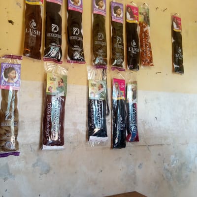 The image depicts a wall display of packaged hair extensions in a store, likely a beauty supply shop, in Nsugbe, Nigeria. The wall is painted a light yellow color and has visible imperfections. A variety of hair extension brands, including "LUSH Hair," "Darling," and "X-Pression" are displayed in their original packaging. Each package contains a sample of the hair extensions inside, as well as the brand logo and a woman's portrait. There are a range of hair colors, including black, brown, and blonde. The extensions are attached to the wall by nails. The floor is brown and partially visible at the bottom of the image.