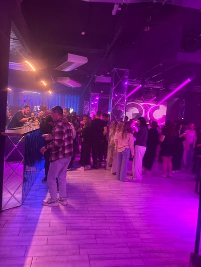 The image depicts the interior of a nightclub, with a focus on a group of people gathered near a bar and on the dance floor, illuminated by vibrant purple and pink lighting. The environment appears to be a trendy establishment, with modern decor including illuminated vertical panels and a large screen displaying a logo in the background. The floor is tiled and reflects the colorful lighting.

In the foreground, a man in a plaid shirt and jeans stands facing away from the camera, with his left hand near his face. To his right, another person in a black top and white pants is partially visible. To the left of the foreground subject, a bar area is visible with a bartender and several patrons. The bar is well-lit and stocked with bottles.

Further into the background, more people are seen mingling, dancing, and standing in groups. The lighting creates a dynamic and energetic atmosphere typical of a nightclub.

There is no information within the image itself to suggest the location is Kirov, Russia.