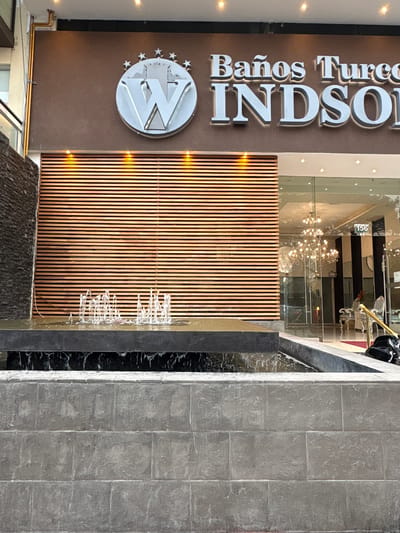 This image captures the exterior of a commercial establishment, specifically "Baños Turcos WINDSOR" (partially visible), located in San Isidro, Peru. In the foreground, a multi-tiered fountain is present, featuring a base constructed from rectangular light grey, textured stone blocks. Water cascades from a higher, dark grey basin into a lower basin with a black marbled interior, where multiple active water jets spray upwards.
To the left, a vertical wall section is clad in dark, stacked stone. Behind the main fountain basin, a large wall area is covered with horizontal wooden slats, illuminated by three recessed spotlights from above. Above this wooden section, two brass-colored pipes run along and up a brown-painted facade.
The upper part of the brown facade displays the establishment's signage. On the left is a silver circular emblem featuring a stylized "W" crowned by a smaller structure and surrounded by five small stars. To its right, raised silver letters spell "Baños Turcos" and "WINDSO" (the 'R' is cropped from view).
To the right of the wooden slat wall, the building's main entrance is a glass facade, reflecting and revealing the interior. Through the glass, an elegant setting is visible, including multiple ornate chandeliers, classical-style chairs with high backs, and vertical black and white striped wall decor. A small white sign with the number "156" in black is affixed to the glass near the upper right of the entrance. A golden handrail guides towards the entry, and a black bag is resting on the ground nearby. No individuals are visible in the scene.