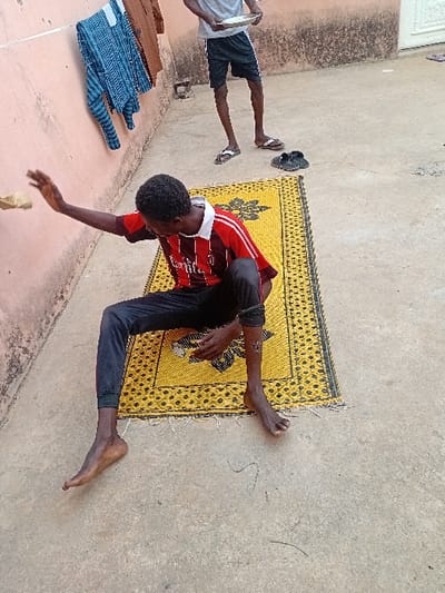 An outdoor scene in Ayama, Nigeria, features two males on a concrete surface against a light pink wall. In the foreground, a dark-complexioned male sits barefoot on a yellow woven mat with black geometric patterns. He wears a red and black striped short-sleeve shirt and black pants, with an apparent mark or injury on his right lower leg. His right arm is raised, hand open, seemingly interacting with a light-colored, indistinct object held near his hand. Behind him, to the right, a second dark-complexioned male stands, wearing a light grey t-shirt, dark shorts with white side stripes, and white flip-flops. He holds a light-colored shallow bowl or plate in both hands. Near his feet on the concrete are two dark shoes or slippers. On the pink wall to the left, a blue and white patterned long-sleeve shirt and a brown garment hang. A white door is partially visible in the far background. The ground is concrete with minor debris.