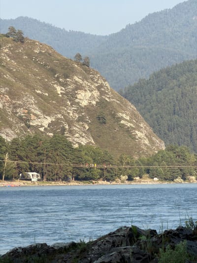 The image shows a wide river in the foreground, with choppy water reflecting the sky. In the middle ground, a dense line of evergreen trees runs along the riverbank, backed by a steep, rocky mountainside dotted with sparse vegetation and a few trees. A suspension bridge spans the river, with several people visible walking across it. To the left of the bridge on the riverbank is a white, tent-like structure. In the background, rolling, forest-covered hills recede into a hazy distance. The scene appears to be set in a natural, mountainous landscape.