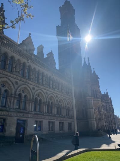 The image shows the Bradford City Hall, located in Bradford, United Kingdom at latitude 53.793, longitude -1.753. The sun is shining brightly behind a tall tower, casting a glare on the stone building. The building has a long facade with many arches, and a single person is walking in front of it. The sky is blue and cloudless. The grass is green and lush.