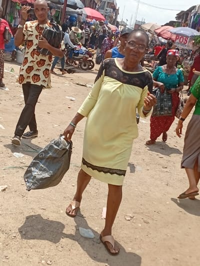 The image captures a vibrant and bustling outdoor market or street scene, likely under bright daylight. The ground is unpaved, appearing dusty and sandy, with some scattered debris like small white pieces of plastic or paper.
In the immediate foreground and center, a woman is walking towards the viewer, looking directly at the camera. She is wearing a light yellow, knee-length dress with a boat-neckline, featuring black lace detailing across the chest and a black lace trim near the hem. She has short, dark hair, wears glasses, and is adorned with a green bracelet on her left wrist. She is wearing light-colored, possibly beige, flip-flops or open-toed sandals. In her left hand, she carries a large, dark grey or black bag, which swings slightly, indicating her movement. Her right hand is slightly raised, palm facing outwards.
To the left of the main subject, slightly behind her, a man with a bald head is also walking. He is wearing a distinctive light-colored, long-sleeved shirt patterned with numerous brown lion heads. He wears dark-colored pants and dark shoes. He carries a black bag tucked under his left arm. Like the woman, he appears to be looking towards the camera.
The background is filled with numerous people and market activity. There are several market stalls visible, characterized by their colorful awnings or umbrellas in shades of red, brown, and blue, suggesting various vendors and goods. To the right of the main woman, another woman in a teal top and a red patterned skirt is walking, carrying a black and white patterned bag. Further to the right, only the lower half of another person in a green top and grey skirt is visible.
In the mid-ground, a motorcycle loaded with goods is parked or moving slowly. Many other pedestrians are present, some identifiable by their clothing, such as a person wearing a straw hat. The lighting is bright, casting distinct shadows on the ground, suggesting it is midday or early afternoon. A white sign with black text is visible in the upper right background, clearly displaying "POS" with "AVAILABLE HERE" written below it. The overall impression is one of a lively, crowded marketplace in full swing.