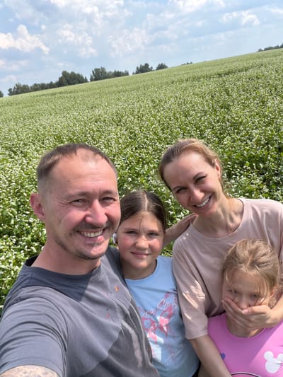 In Nalobikha, Russia, a family of four poses for a selfie in a field of white flowers under a partly cloudy sky. The father, on the left, wears a grey t-shirt and smiles widely. Next to him is his daughter, wearing a light blue t-shirt. Behind them, the mother, in a light pink t-shirt, smiles and rests her hand on her other daughter's shoulder. This younger daughter, on the right, is wearing a bright pink t-shirt and covers her mouth with her hand. The field of flowers stretches out behind the family, leading to a tree line in the distance.