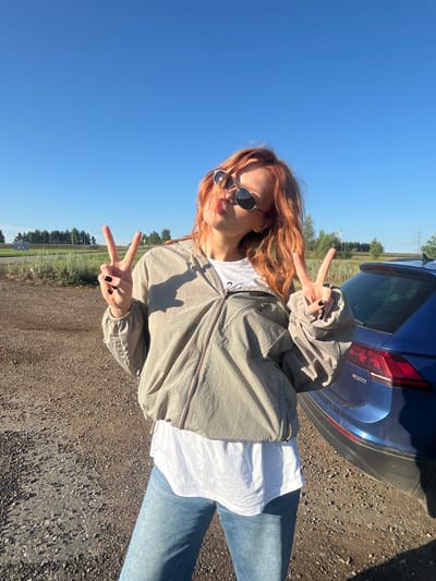 A young woman with auburn hair, wearing sunglasses, a light-colored windbreaker over a white t-shirt, and blue jeans, stands outdoors next to a blue car. She is making a peace sign with both hands. The ground is covered in gravel, and the background shows a clear blue sky, some trees, and distant infrastructure. The location is identified as Verkhnii Sekines', Russia, a rural area, likely on the outskirts of the settlement, indicated by the presence of a dirt road, sparse vegetation, and distant utility poles and transmission towers visible in the background. The lighting suggests it is a sunny day.