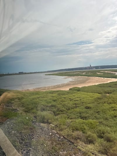 The image depicts a vast landscape featuring a body of water, lush green vegetation, and a distant industrial structure. The water occupies a significant portion of the frame, reflecting the overcast sky. To the right of the water, a dense expanse of green grass stretches into the distance, with patches of brown earth visible in between. The terrain appears somewhat rugged, with scattered rocks in the foreground.
In the far distance, on the left side of the image, an industrial complex is visible. It includes several tall structures, one of which resembles a smokestack or cooling tower, and other buildings that suggest a manufacturing or power generation facility. The sky is predominantly covered with clouds, indicating a possibly cool or overcast day. The overall impression is of a wide, natural environment juxtaposed with industrial development. The image seems to be taken from an elevated vantage point, possibly a bridge or a hillside, looking out over the scene. There is no text visible in the image.