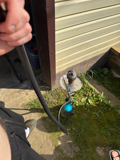 A person's hand holds a black hose, seemingly connected to a hookah. The hookah itself, with a blue glass base, rests on a patch of green grass next to a concrete surface. Above the grass, a light-colored wooden-paneled wall is visible, with a dark brown vertical beam running down the left side. The ground is a mix of dirt, grass, and concrete. The scene appears to be outdoors, possibly in a yard or garden area. There are no other people or animals visible in the frame. The lighting suggests it is daytime.