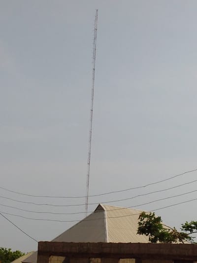 The image displays a tall, slender, reddish-brown lattice telecommunication tower positioned vertically against a light, uniform grey sky in Ajaokuta, Nigeria. The tower extends from the mid-frame to near the top edge. In the lower-midground, five dark, parallel utility lines span horizontally across the scene. Below these lines, the upper section of a building is visible, featuring a light-beige corrugated metal hip roof with sharp angles. Beneath the roof, a section of an unplastered reddish-brown brick wall is apparent. Green foliage is present to the right of the building's roof and faintly in the lower-left corner. No individuals or specific activities are depicted.