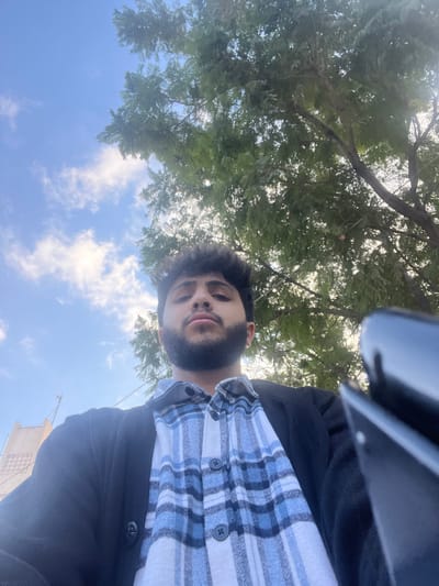 A man with dark hair and a beard looks up at the camera from a low angle. He wears a black jacket over a blue and white plaid shirt. Behind him, a blue sky dotted with clouds peeks through the dense green leaves of a tree. Part of a black device is visible in the top right corner, suggesting the photo was taken with a phone. The bottom of a light-colored building is faintly visible.