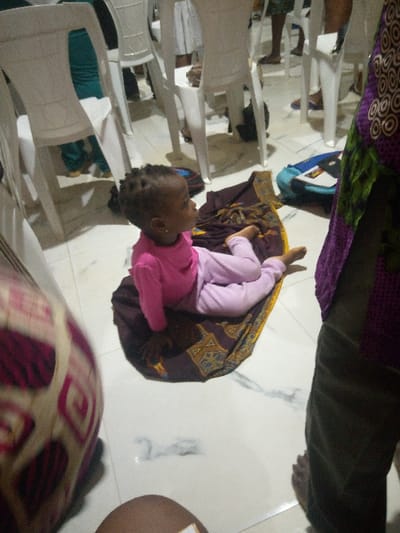 A toddler with braided hair, wearing a pink top and leggings, sits on a patterned fabric square on a white tiled floor. The child looks to the right, away from the camera. White plastic chairs surround the scene. A person stands to the right wearing a patterned purple and green fabric. Part of another person wearing a patterned fabric can be seen to the left. A blue bag is visible in the background.