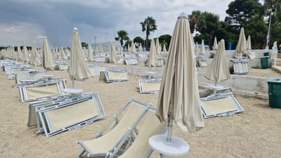 The image depicts a beach with numerous folded beach chairs and closed umbrellas arranged on the sand. The scene is set on a beach, likely in Taranto, Italy, indicated by the location context. The sky is overcast and cloudy, suggesting a potentially stormy or at least a dull day. Several palm trees are visible in the background, along with other vegetation, and a white concrete structure runs along the back of the beach, supporting some of the umbrellas and chairs. Two green trash bins are also visible on the right side of the image. The chairs are made of a light-colored fabric stretched over a white frame, and the umbrellas are a light beige color. Some chairs are stacked, while others are placed individually. There are no people visible in the scene. The overall impression is one of a beach that is either closed for the season, preparing for bad weather, or simply not yet open for the day. The arrangement of the chairs and umbrellas suggests a structured setting, likely a commercial beach resort.