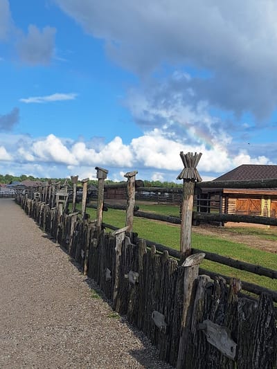 A rustic, wooden fence, made of rough-hewn logs and branches, runs diagonally from the lower left corner to the upper right of the image. The fence is old and weathered, with some sections appearing to be constructed from tree stumps and bark. Small, wooden birdhouses or feeders are attached to some of the fence posts. Beyond the fence, a grassy field stretches out, dotted with more wooden structures and fences. In the background, a cluster of buildings, likely farm buildings or rustic cabins, are visible, with a dark, shingled roof on one of them. The sky above is a bright blue, with fluffy white clouds and a faint rainbow arcing across the upper right portion of the frame. The ground in the foreground is a gravel path. The scene depicts a rural or historical setting, possibly an open-air museum or a farmstead, in the Malakhovetsky Selsoviet, Belarus.