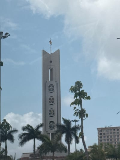 The image displays a tall, slender, grey concrete monument tower, identified as a memorial, located within Hòa Cường Ward, Vietnam, beneath a partly cloudy blue sky. The tower's apex is pointed, crowned with a golden star emblem. Below this, a small circular opening and a longer vertical rectangular slit are visible. The main shaft of the tower features four uniformly spaced, recessed square panels, each containing an obscured bas-relief. The lower section of the tower is partially concealed by foreground and midground tropical vegetation, consisting of multiple palm trees with dark green fronds and at least one tree with lighter green foliage. A metallic utility pole with multiple light fixtures is partially visible on the far left. In the distant background on the right, a multi-story building with a grid of windows is partially discernible, suggesting an urban setting. No individuals or specific activities are present in the scene.