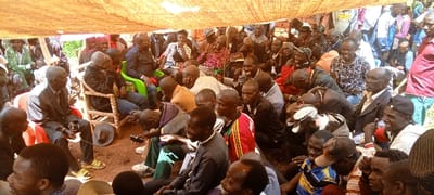 A large group of people, mostly men, are gathered under a makeshift tent. Many are seated on chairs or the ground, attentively listening. Individuals are dressed in a mix of formal attire, casual clothing, and traditional wear. The setting appears to be outdoors with a reddish-brown ground and some greenery visible in the background. The overall atmosphere is one of community engagement or a formal gathering.