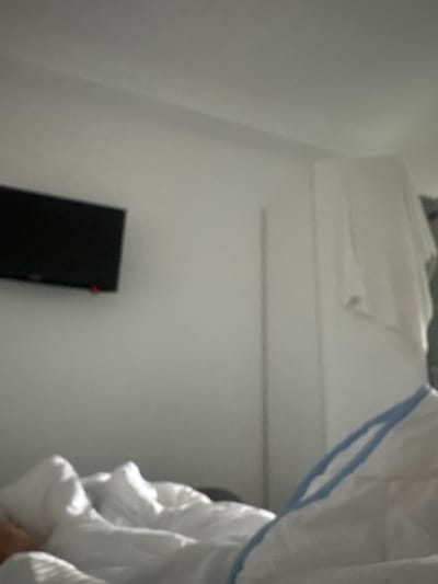 The image depicts an indoor scene, likely a bedroom, with a focus on a person covered by white bedding. A black television is mounted on a white wall to the left of the frame. A small red light is visible on the television, suggesting it is powered on or in standby mode. To the right, a white cloth hangs from a tall structure, possibly a closet or wardrobe. The overall environment appears to be a living space, possibly a hotel room or a domestic bedroom in Stațiunea Zoologică Marină Agigea, Romania. The person in the bed is mostly obscured by the covers, with only a small portion of their arm visible. The lighting suggests it is daytime, with natural light entering the room.