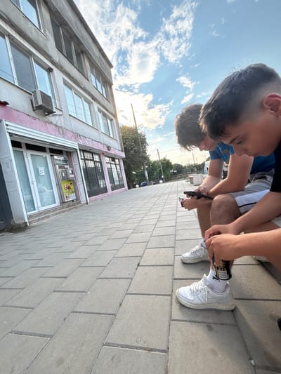 The image shows two young males sitting on a sidewalk in front of a building in Bulgarovo, Bulgaria. The building appears to be a commercial establishment with multiple windows and an air conditioning unit visible. The sidewalk is paved with rectangular concrete slabs. One of the males is looking at his phone, while the other is fiddling with a can. Both are wearing athletic shoes. The sky is partly cloudy with sunlight visible. The overall scene depicts a casual moment outdoors on a sunny day.