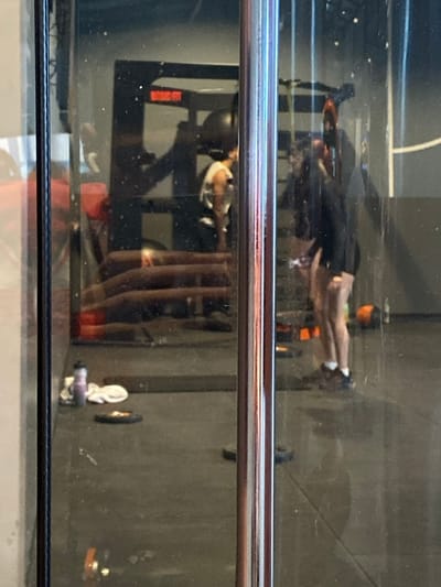 The image depicts an interior view of a Basic-Fit gym located in Hœnheim, France, captured through a reflective glass surface. A vertical, chrome-plated metal bar occupies the central foreground, partially obstructing the view.
Within the gym, two individuals are visible, likely reflected in the glass. On the left side of the reflection, a person wearing a white sleeveless top is partially seen from behind, standing near a black gym rack which bears the "BASIC-FIT" logo in red lettering. Below this, several horizontal, reddish-brown upholstered benches or steps are stacked on the dark floor.
On the right side of the reflection, another individual is standing, clad in a dark long-sleeved top and dark shorts, exposing their legs. They wear athletic shoes with white socks and are positioned facing left. On the dark gym floor in the foreground, near the left edge, a light purple water bottle rests beside a white towel and a dark weight plate. Another dark weight plate is visible further right on the floor, and a black spherical object, possibly a medicine ball, is on the floor near the right-side individual's feet. The walls are dark gray. The scene appears well-lit by interior lighting.