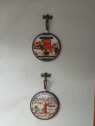 Two round, decorative wall hangings are displayed vertically on a textured, light gray wall. Each hanging is suspended from a metal dragonfly-shaped hook by a dark braided cord.
The upper hanging features a scene with the text "WABASH DEPOT" at the top. The artwork depicts a red arched doorway, a shelf with various items including baskets of fruit and gifts, and a woman standing with a doll. A spoked wheel is also visible. Below the image, the text reads, "His eyes upon you as you come."
The lower hanging is also circular and has text around its perimeter, including "A, B, C, D, E, F, G, H, I, J, K, L, M, N, O, P, Q, R, S." In the center of this hanging is a depiction of a tree with children playing in and around it, including on a swing. Text at the bottom reads, "OUR ROOTS GROW SOIL OF GOLD."
The wall has a consistent, ridged texture. The lighting appears to be from above and to the right, casting subtle shadows. There is a vertical wooden trim piece visible on the far right edge of the frame. The scene is static and shows no signs of activity. The location is unknown.