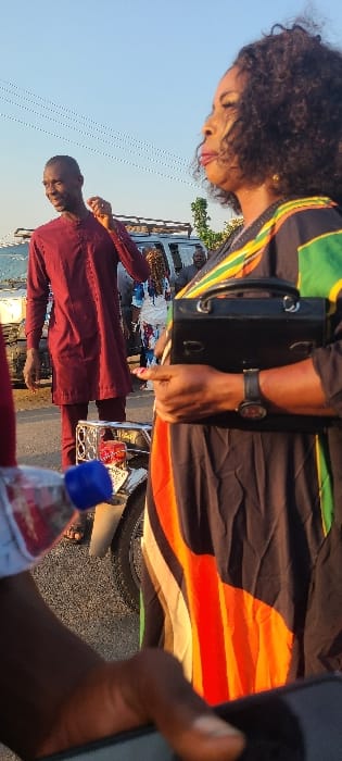 Two people stand outside on a sunny day. A woman with dark, curly hair wears a colorful dress and carries a black handbag. Beside her, a man in a maroon shirt stands in front of a parked vehicle. A water bottle rests on a nearby surface. A hand holding a phone is in the bottom right corner of the frame. The image is rotated clockwise.