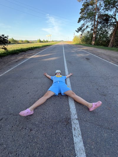 A person wearing a light blue t-shirt and shorts, a white baseball cap, and pink Crocs lies spread-eagled on a gray asphalt road with a white lane divider. The road is surrounded by green foliage and a blue sky. The location is not apparent from the given image.