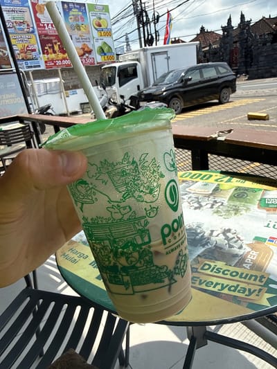 A person is holding a disposable Polos coffee cup filled with iced coffee on a table in Kutuh, Indonesia. The coffee cup has a green design on it and a straw. The table has a circular ad for the Aper discount. A black metal chair is visible. In the background, there are billboards advertising food items and prices, as well as a black car and a white delivery truck parked on the street. Power lines and buildings are also visible.