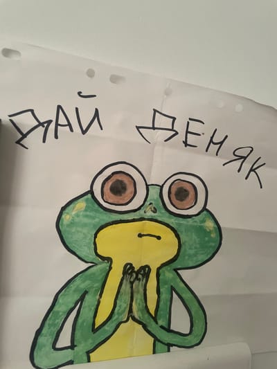 The image shows a drawing of a frog with the Russian text "ДАЙ ДЕНЯК" written above it. The text translates to "Give money." The frog is green with yellow coloring around its mouth and a white background. The drawing appears to be made with markers and is on a piece of paper with punched holes. There is no visible indication of this image being located in Vénissieux, France, or any other specific location.