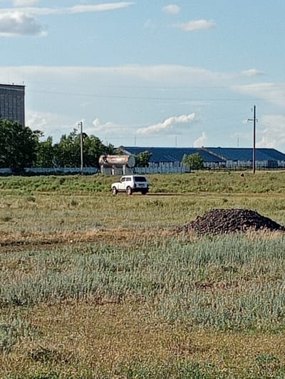 In a wide shot of a rural or industrial area, a white SUV is parked on a dirt road in a field. Behind the SUV, a large, long building with a blue roof is visible, bordered by a white fence. To the left of this building, a tall, dark-colored industrial structure stands, partially obscured by green trees. A large cylindrical tank sits in front of the trees. The foreground consists of a grassy field with a pile of dark material in the lower right corner. A single utility pole is visible to the right of the blue-roofed building. The sky above is partly cloudy. The location is described as Oktyabrskoye, Kazakhstan.